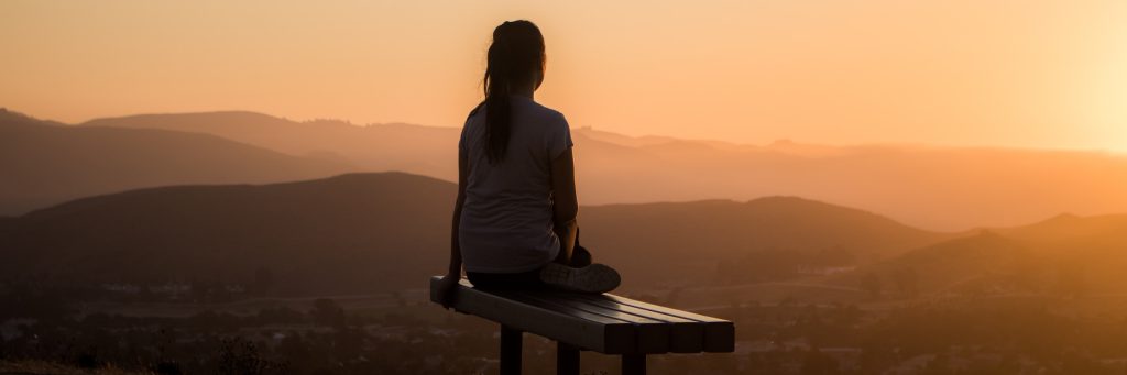 Die Rückansicht eines Mädchens bei einem Sonnenuntergang. Sie sitzt auf einer Bank hoch über einer bergigen Landschaft
