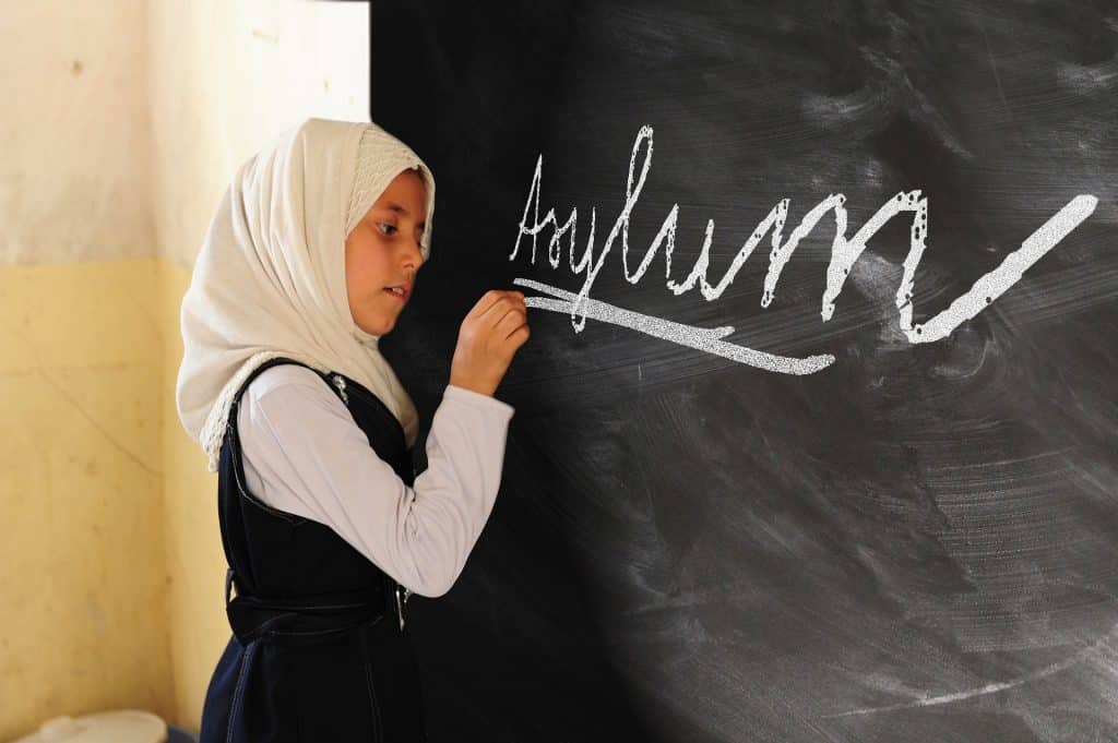 Schülerin mit einem weißen Kopftuch vor einer Schultafel, auf die sie das Word "Asylum" geschrieben hat